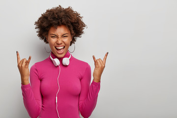 Energetic curly female meloman makes rock n roll gesture, has joyous rebellious expression, wears pink turtleneck, headphones and earrings, isolated over white background, blank space aside.