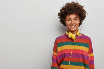 Energetic enthusiastic woman with curly crisp hair, listens music via headset, smiles broadly,...