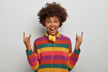 Dark skinned curly woman shows rock n roll gesture, enjoys heavy metal, has overjoyed expression, wears striped sweater, looks gladfully at camera, models against white background. Rock this party