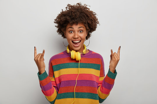 Dark skinned curly woman shows rock n roll gesture, enjoys heavy metal, has overjoyed expression, wears striped sweater, looks gladfully at camera, models against white background. Rock this party