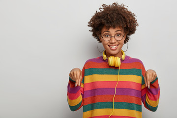 I found what you searched. Good looking ethnic female points index fingers down, shares nice offer, feels glad, wears spectacles and sweater, isolated over white background. Advertising concept