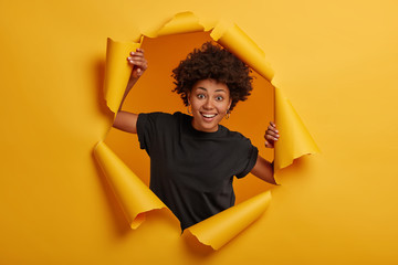 Photo of cheerful dark skinned girl wears black t shirt, stands in torn paper wall, looks directly...