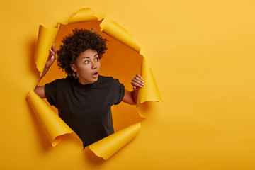 Fototapeta Photo of surprised Afro woman in black t shirt gasps from amazement, looks with scared face expression aside, dressed in black t shirt, unexpected to see something horrible, poses in torn paper wall obraz