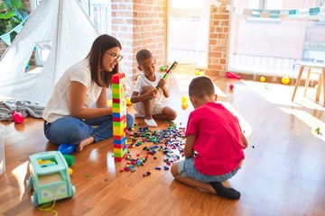 Young beautiful teacher and toddlers playing with building blocks around lots of toys at kindergarten