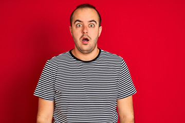 Young man wearing striped navy t-shirt standing over isolated red background afraid and shocked with surprise expression, fear and excited face.