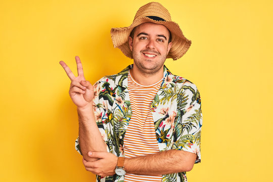Young man on vacation wearing summer shirt and hat over isolated yellow background smiling with happy face winking at the camera doing victory sign. Number two.