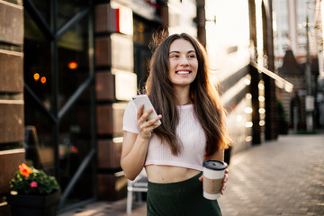 Smiling woman using phone and drink coffee on the street in summer day.