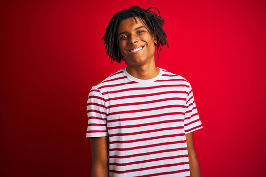 Young afro man with dreadlocks wearing striped t-shirt standing over isolated red background with a happy and cool smile on face. Lucky person.