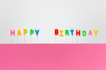 Happy Birthday greeting card with wax colorful letters candles on white pink paper background. Photo with copy blank space.