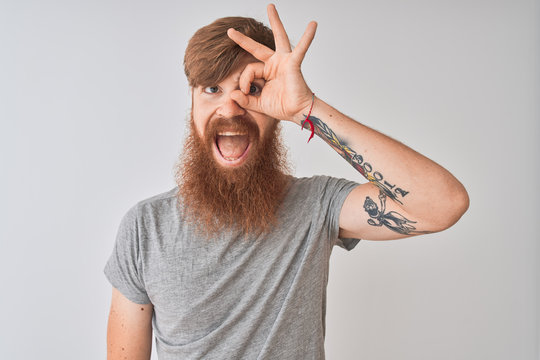 Young redhead irish man wearing t-shirt standing over isolated grey background doing ok gesture with hand smiling, eye looking through fingers with happy face.