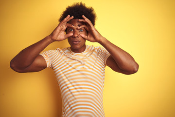 American man with afro hair wearing striped t-shirt standing over isolated yellow background Trying to open eyes with fingers, sleepy and tired for morning fatigue