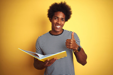 Afro american student man reading book standing over isolated yellow background happy with big smile doing ok sign, thumb up with fingers, excellent sign