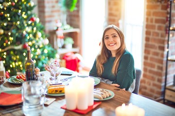 Young beautiful woman sitting eating food around christmas tree at home happy face smiling with crossed arms looking at the camera. Positive person.