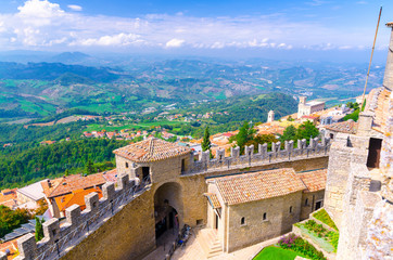 Fototapeta na wymiar Aerial top panoramic view landscape with valley, green hills, fields, villages of Republic San Marino suburban district, blue sky white clouds background and stone fortress wall and tower with merlons