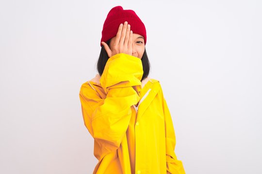Young beautiful chinese woman wearing raincoat and wool cap over isolated white background covering one eye with hand, confident smile on face and surprise emotion.