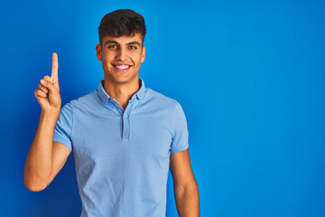 Young indian man wearing casual polo standing over isolated blue background showing and pointing up with finger number one while smiling confident and happy.