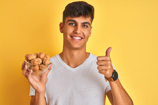 Young indian man holding bowl with walnuts standing over isolated yellow background happy with big smile doing ok sign, thumb up with fingers, excellent sign