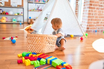 Adorable toddler sitting on the floor playing with wicker basket and balls around lots of toys at kindergarten