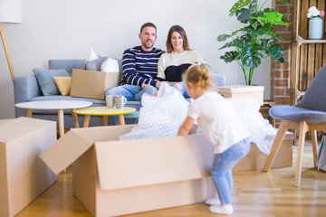 Beautiful family, parents sitting on the sofa drinking coffee looking his kid playing at new home around cardboard boxes