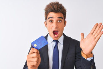 Young handsome businessman wearing suit holding credit card over isolated white background very happy and excited, winner expression celebrating victory screaming with big smile and raised hands