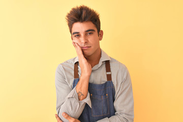 Young handsome employee man wearing apron standing over isolated yellow background thinking looking tired and bored with depression problems with crossed arms.