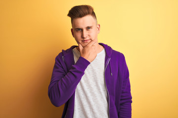 Young handsome man wearing purple sweatshirt standing over isolated yellow background looking confident at the camera with smile with crossed arms and hand raised on chin. Thinking positive.