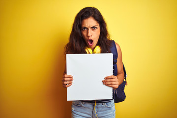 Obraz na płótnie Canvas Young beautiful student woman holding banner standing over isolated yellow background scared in shock with a surprise face, afraid and excited with fear expression