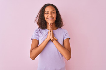 Young brazilian woman wearing t-shirt standing over isolated pink background praying with hands...