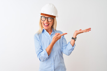 Middle age architect woman wearing glasses and helmet over isolated white background amazed and smiling to the camera while presenting with hand and pointing with finger.