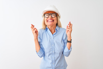 Middle age architect woman wearing glasses and helmet over isolated white background gesturing finger crossed smiling with hope and eyes closed. Luck and superstitious concept.