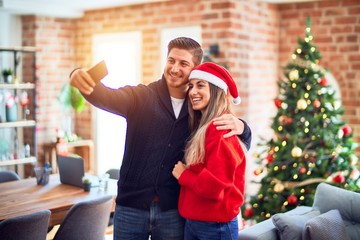 Young beautiful couple smiling happy and confident. Standing and hugging make selfie by camera around christmas tree at home