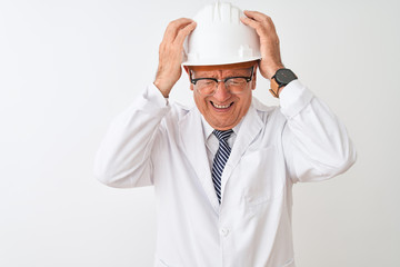 Senior grey-haired engineer man wearing coat and helmet over isolated white background suffering from headache desperate and stressed because pain and migraine. Hands on head.