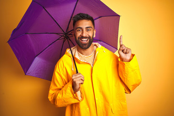 Young indian man wearing raincoat and purple umbrella over isolated yellow background surprised...