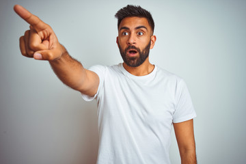 Young indian man wearing t-shirt standing over isolated white background Pointing with finger surprised ahead, open mouth amazed expression, something on the front