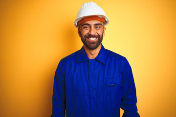 Handsome indian worker man wearing uniform and helmet over isolated yellow background with a happy...