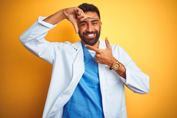 Young indian doctor man standing over isolated yellow background smiling making frame with hands and fingers with happy face. Creativity and photography concept.