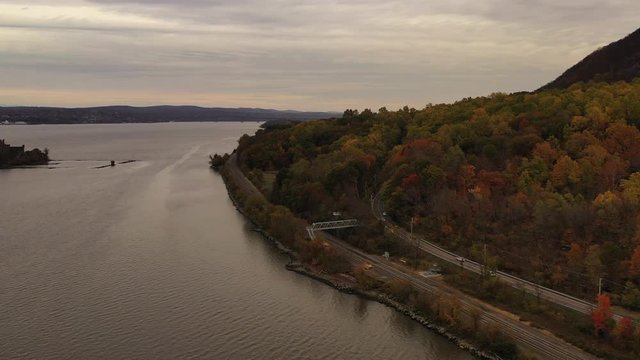aerial drone shot over the Hudson River, while the camera dolly in & descends down, trucks left towards the autumn colored tree tops, a roadway & train tracks on a cloudy evening in Beacon, NY