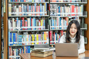 Asian young Student in casual suit doing homework and using technology laptop in library of university or colleage with various book and stationary over the book shelf background, Back to school