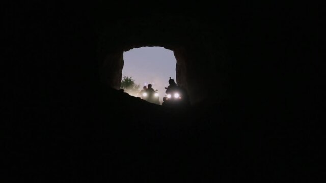 Row of several extreme quadrocycles switched on headlights and with men wearing helmets are actively riding and moving around inside of huge empty dark cave.