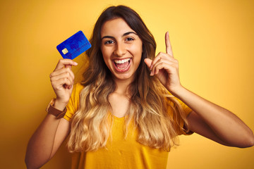 Young beautiful woman holding credit card over yellow isolated background surprised with an idea or...