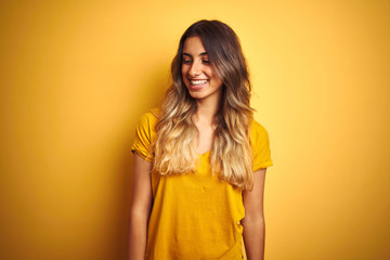 Young beautiful woman wearing t-shirt over yellow isolated background looking away to side with smile on face, natural expression. Laughing confident.