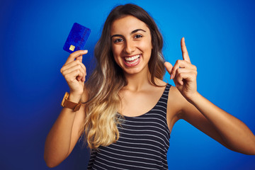 Young beautiful woman holding credit card over blue isolated background surprised with an idea or question pointing finger with happy face, number one