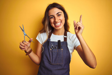 Young beautiful hairdresser woman holding scissors over yellow isolated background very happy...