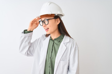 Young chinese engineer woman wearing coat helmet glasses over isolated white background very happy and smiling looking far away with hand over head. Searching concept.