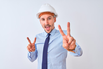 Young business man wearing contractor safety helmet over isolated background smiling looking to the camera showing fingers doing victory sign. Number two.