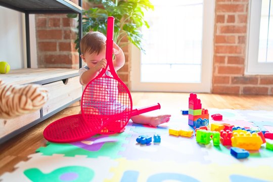Beautiful toddler sitting on puzzle carpet holding tennis racket at kindergarten