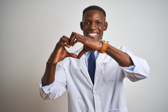 Young african american doctor man wearing coat standing over isolated white background smiling in love doing heart symbol shape with hands. Romantic concept.