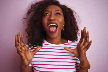 Young african american woman wearing striped t-shirt standing over isolated pink background crazy and mad shouting and yelling with aggressive expression and arms raised. Frustration concept.