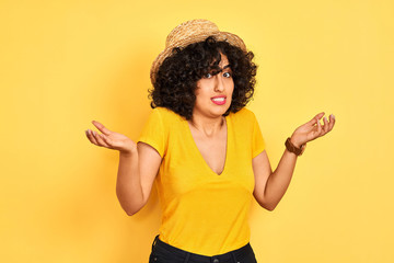 Young arab woman with curly hair wearing t-shirt and hat over isolated yellow background clueless...