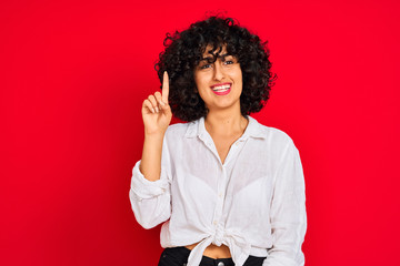 Young arab woman with curly hair wearing white casual shirt over isolated red background showing and pointing up with finger number one while smiling confident and happy.